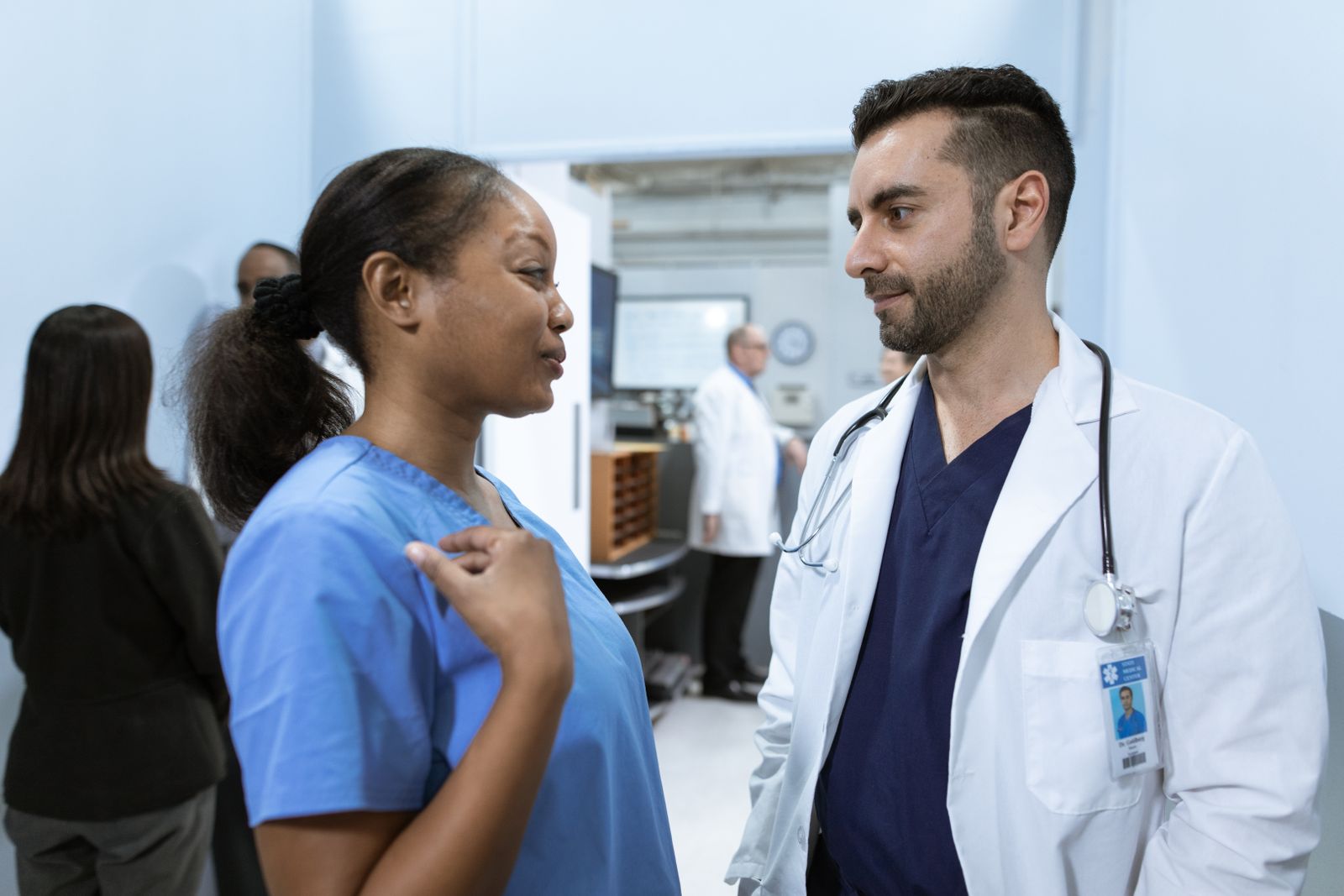 Two health workers in scrubs and a white coat talking in a hospital corridor